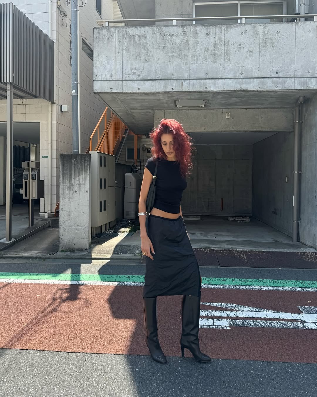 A woman standing wearing a black slip skirt, tall black boots, and a basic black tee.