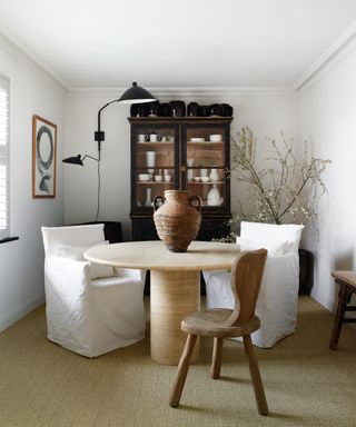 monochrome dining room with round travertine table, two white linen chairs, rustic wooden chair and black cabinet displaying ceramics