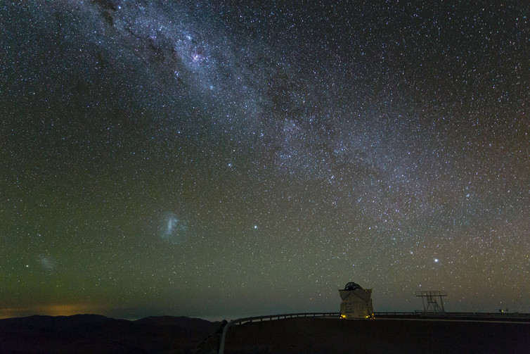 A super starry sky has a glowing streak down the middle and two blurred blobs toward the lower left, near the horizon.