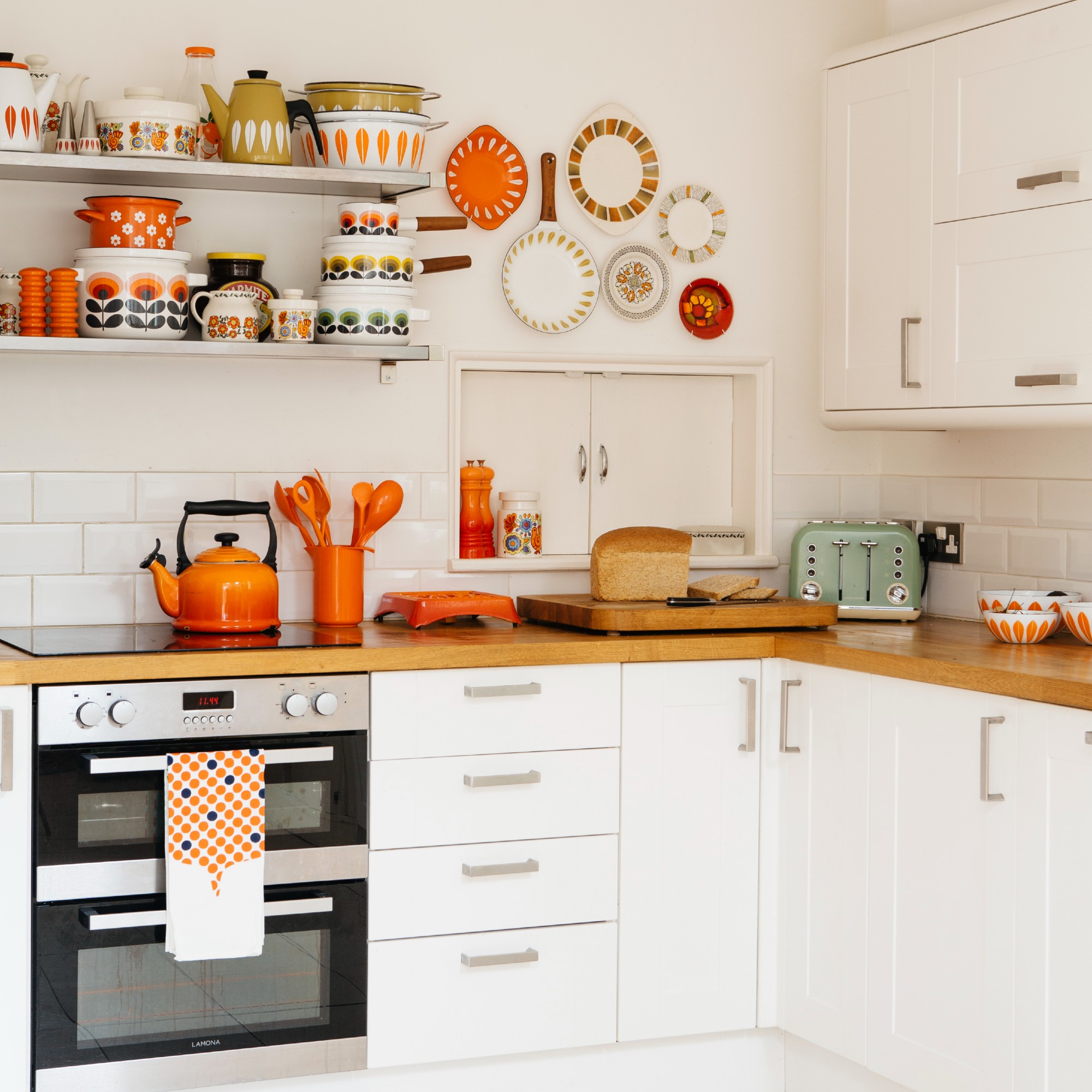 A white kitchen with open shelving displaying patterned cookware and orange accents in the form of decorative crockery and utensils