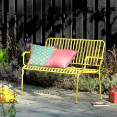 Habitat Indu 2 Seater Metal Garden Bench in Yellow with two colourful garden cushions in a sunny garden 