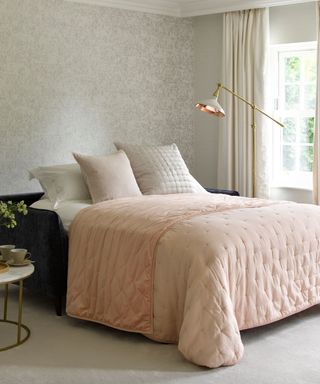 A bedroom with greige walls, a pink bed with light gray throw pillows and a brushed gold standing lamp above it, and a marble round table to the left