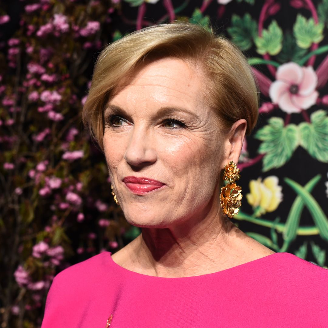 Cecile Richards wears a bright pink dress and stands in front of a wall of flowers