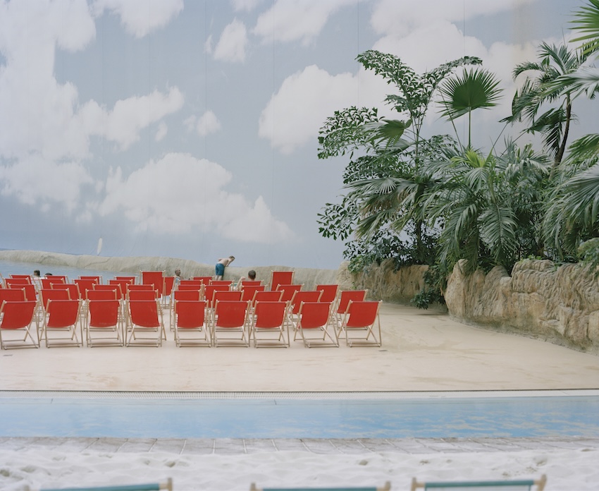 An indoor artificial beach featuring rows of red lounge chairs facing a pool and a large mural of a cloudy blue sky.