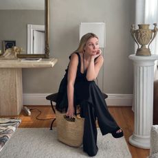 Kerry wears a black top, black satin pants, and a straw tote bag.