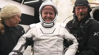 a man in a white spacesuit smiles as he is helped to his feet by two people dressed in black