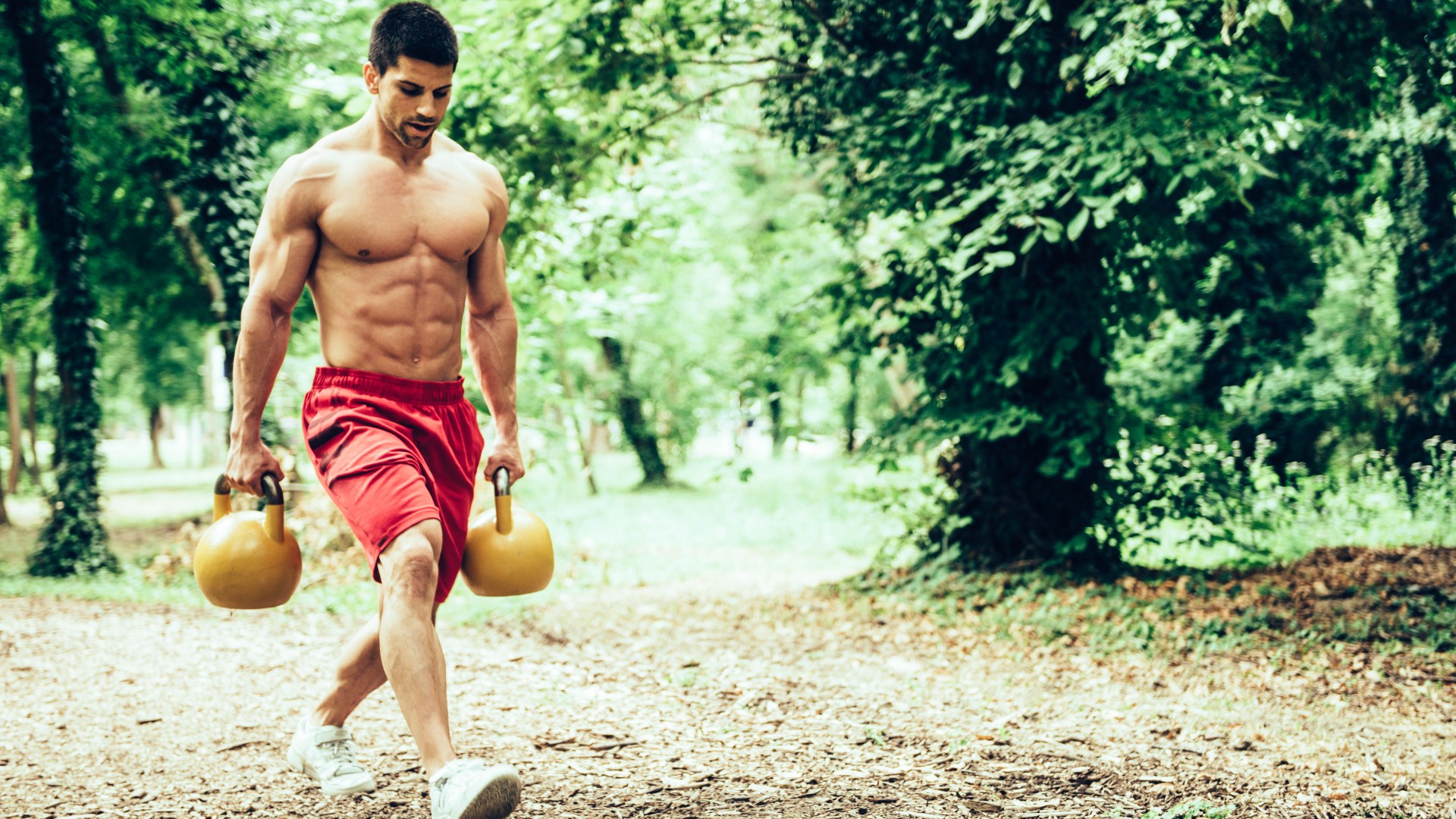 Mann geht mit zwei Kettlebells durch einen Park und macht einen Bauernspaziergang