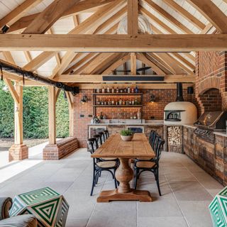 outdoor kitchen under a pergola, wooden beams, wooden cabinetry, pizza oven, BBQ, storage, table and chairs