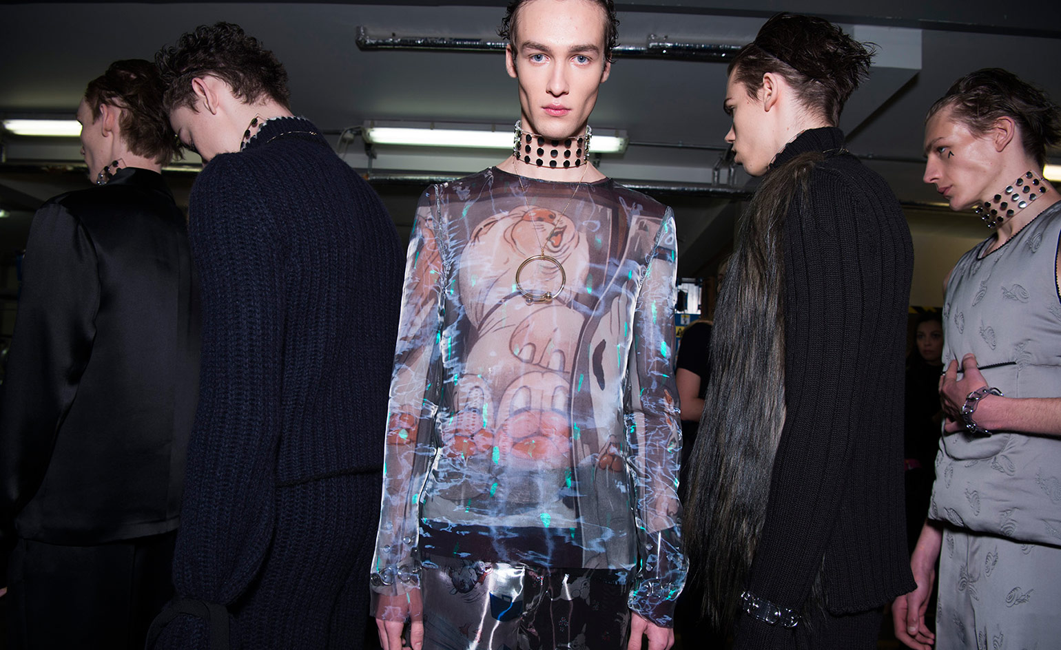 Five male models wearing looks from JW Anderson's collection. Two models are wearing blue ribbed pieces, one with a long hair fur panel at the front. A third model is wearing a dark coloured two piece and a fourth is wearing a grey piece with repeating pattern. The fifth model is wearing a multicoloured top featuring a cartoon-style character and a silver chain with circle pendant. All models are wearing thick chokers with metal studs