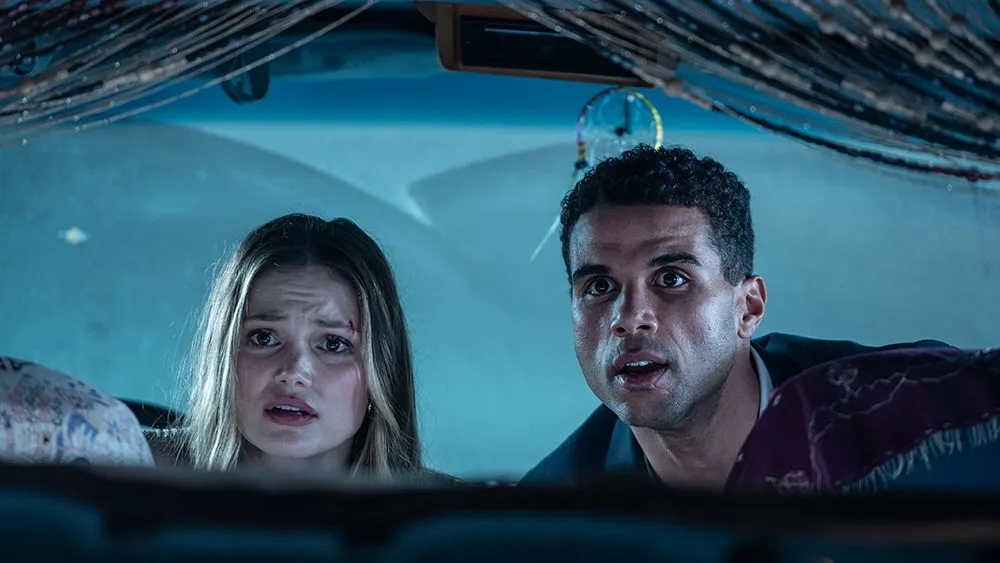 Two people (Olivia Holt as Ally and Mason Gooding as Jay) hide in a room with beaded curtains and a dreamcatcher, in the horror rom-com 'Heart Eyes.'