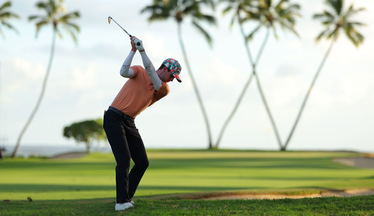 Alex Smalley strikes an iron shot from the rough in front of palm trees which are in the shape of a W