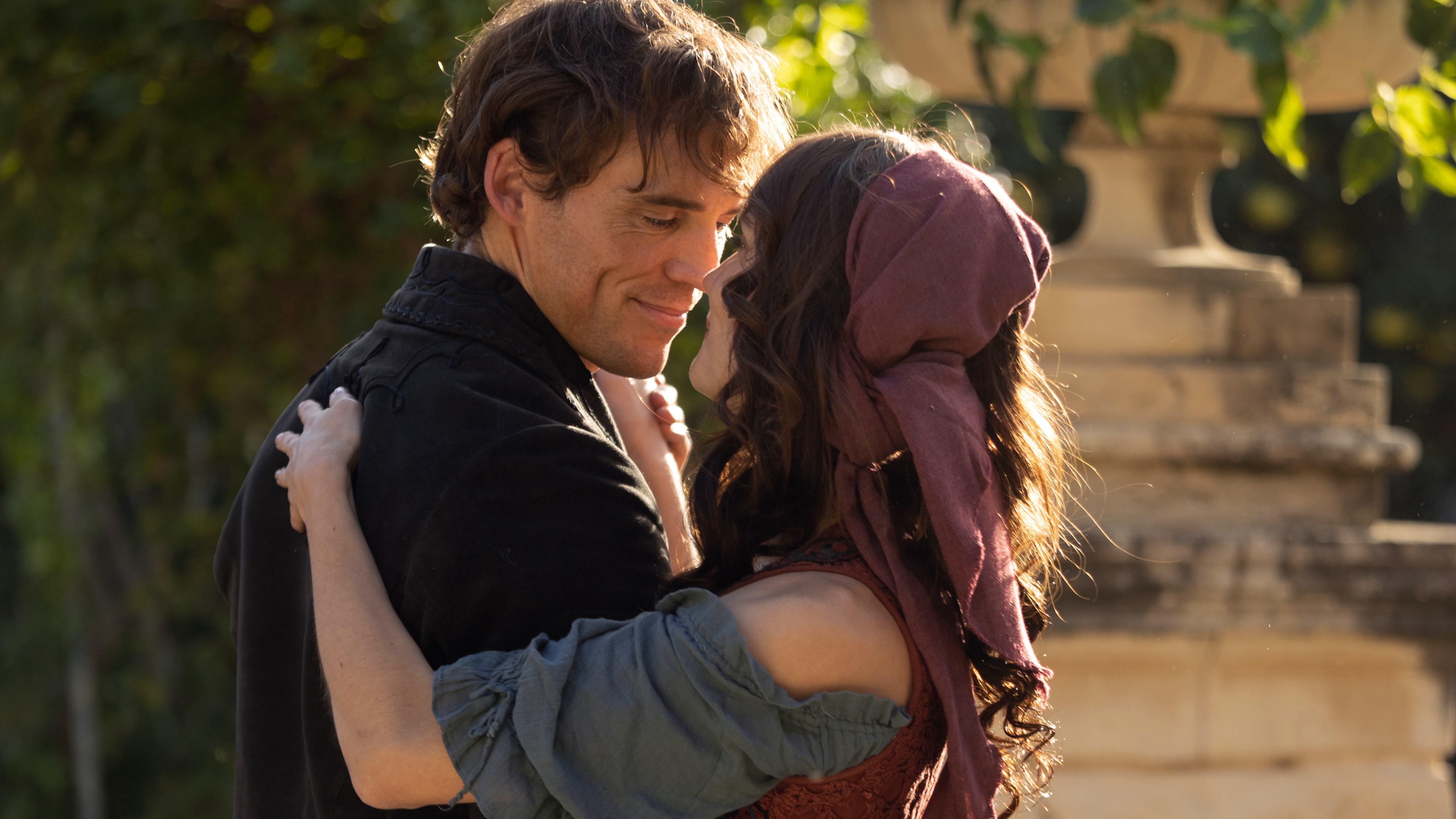 Edmond Dantès (Sam Claflin) and Mercedes (Ana Girardot) are dancing together in a tight embrace, looking happily into each other's eyes. It is a beautiful sunny day, and there are trees and the base of a statue visible behind them.