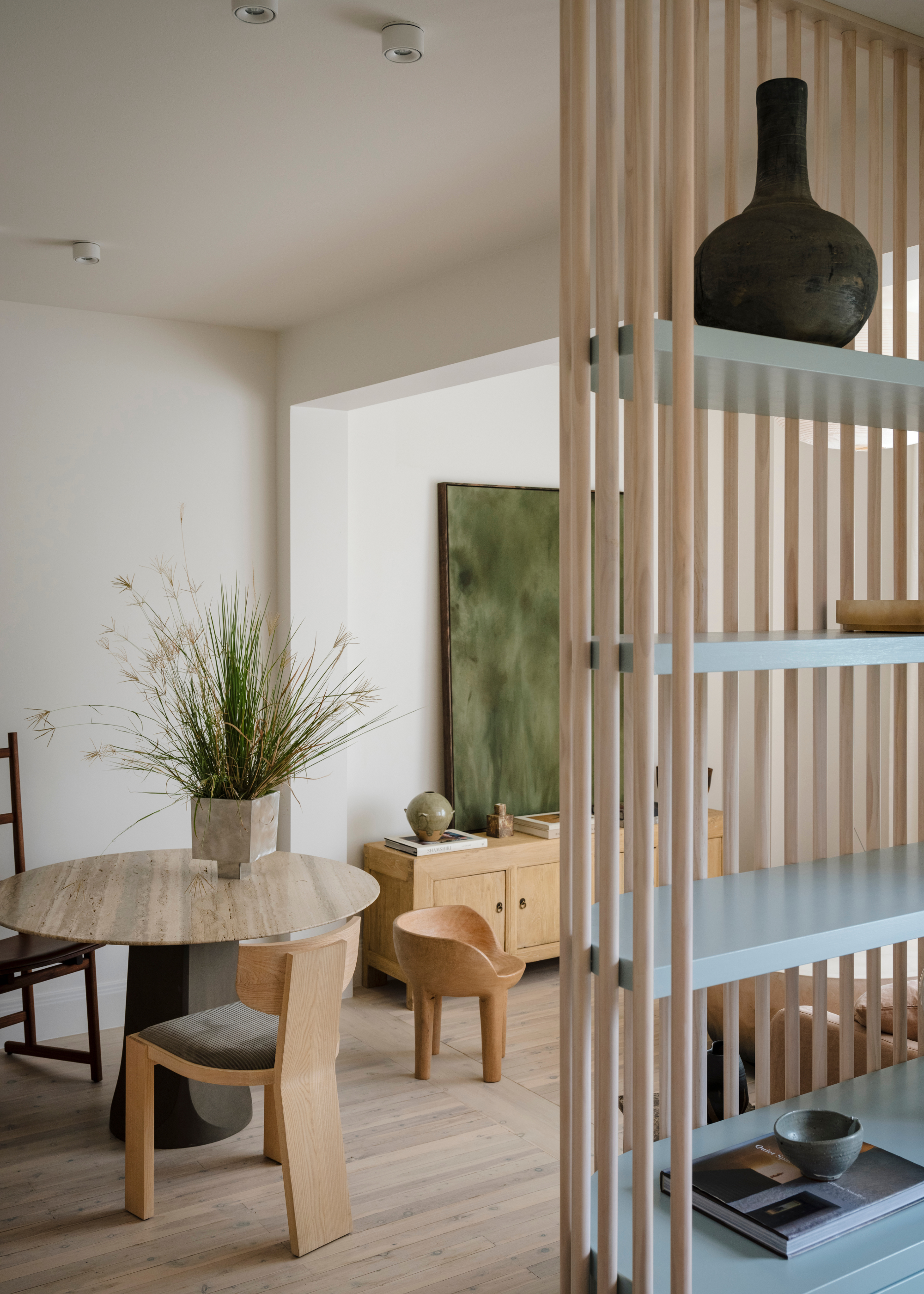 A wood panelled display shelf with a vase and a bowl on a coffee table book. A living room with a small dining table and wooden chairs, a potted plant in a footed planter and a sideboard with a green painting, ceramics and books