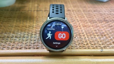 Photo of the Amazfit Active 2 Round smartwatch sitting on a shelf, the display showing an Outdoor Running activity start screen with a "GPS locating" icon, HR icon, and large "GO" button.