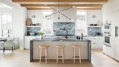 An example of white kitchen backsplash ideas with green marble backsplash and island countertop in a white kitchen with natural wooden ceiling beams and barstools and a statement black metal pendant light.