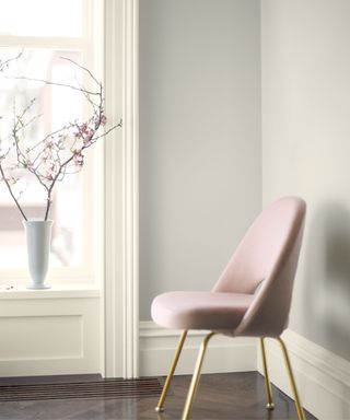 A white room with a light pink curved chair with gold legs, a tall window with a white vase of pink flower branches, and dark brown flooring
