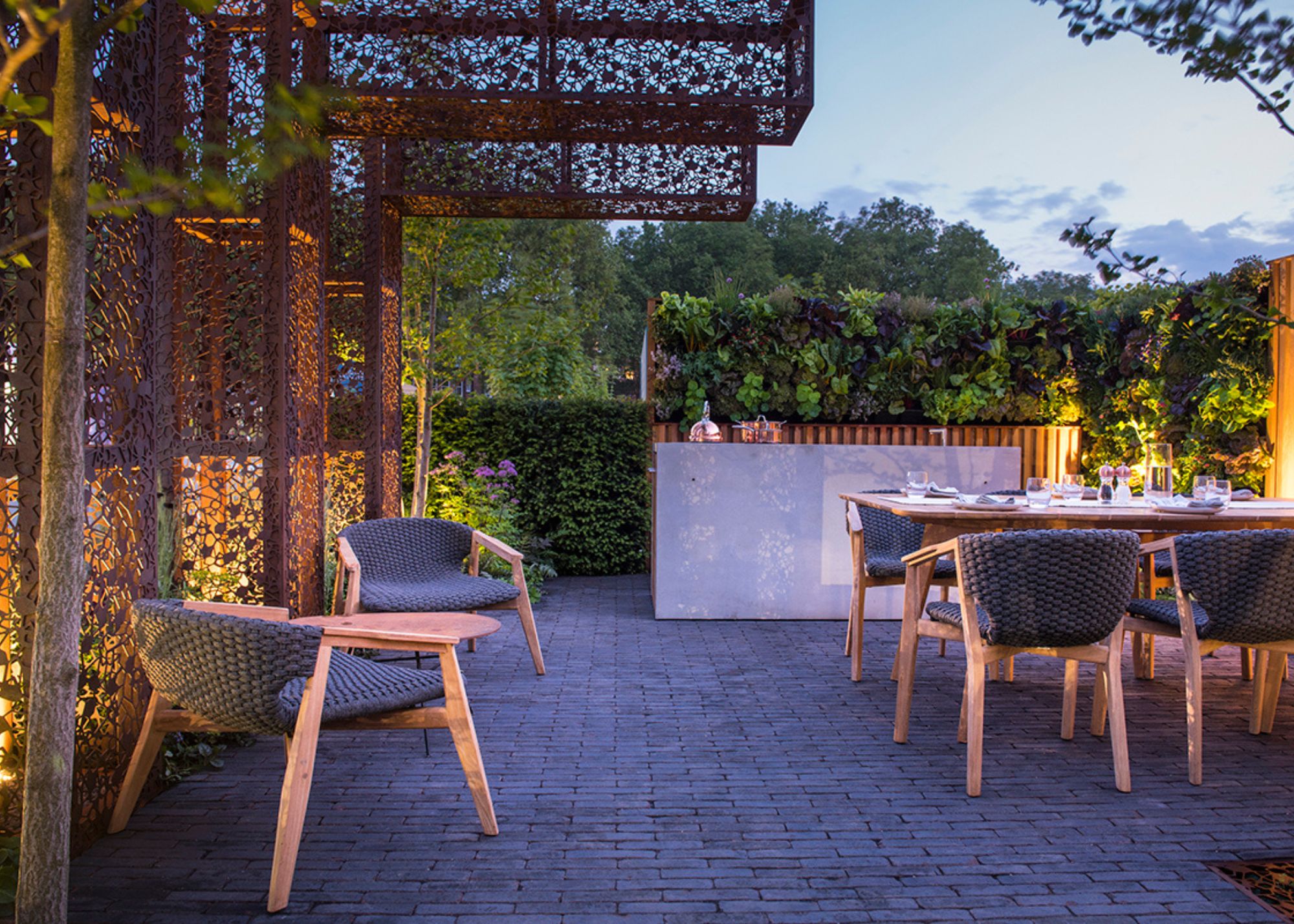 An outdoor area lit along the fence on either side at dusk with a bar/kitchen area at the back a vertical wall of greenery behind, a table and chairs set in front and two chairs along the side wall
