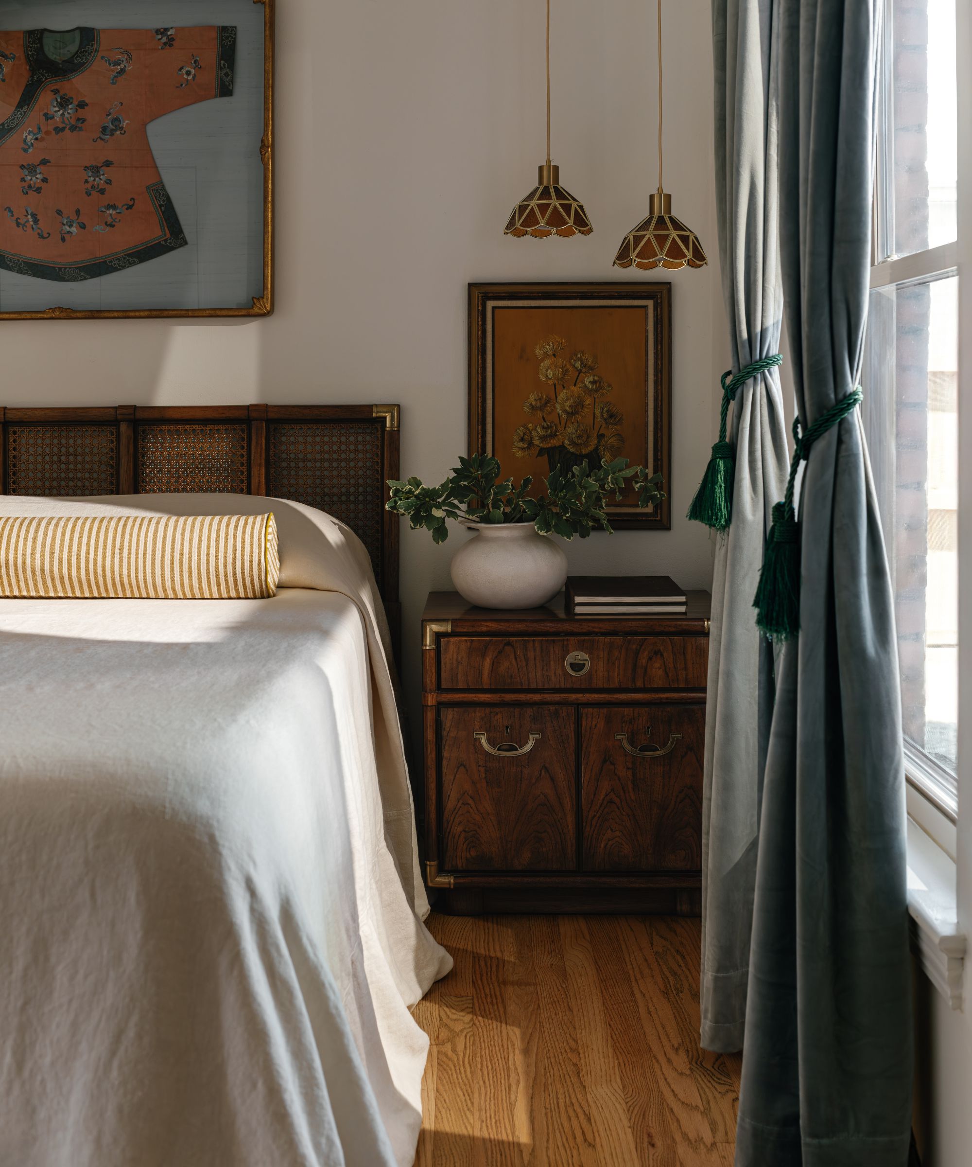 Bedroom with rattan headboard, linen bedding and yellow striped bolster cushion, mahogany sideboard with vase and books, orange painting of flowers, low pendant lights above, and a framed kimono above the bed
