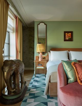 A pastel-shaded hotel room is filled with striking decor elements, including a totemic brass sculpture of an elephant, a geometric sea green rug, a pink velvet banquette, and a pois-relief bedsite table lamp in rose and rattan.