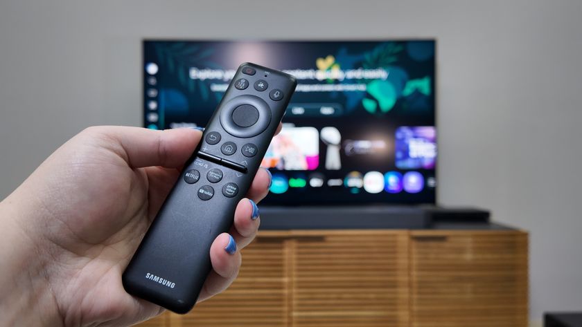 An outstretched hand holds the Samsung Solar Cell remote control with its buttons facing the camera. In the background, the Samsung QN90F is out of focus, displaying its built-in smart platform.