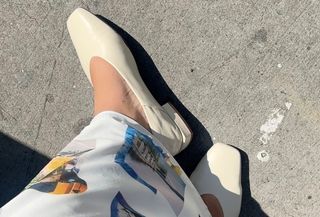 Woman wears white Sam Edelman ballet flats with white printed maxi skirt while standing on street and taking photo from above.