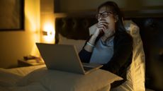 A woman working on her laptop in bed yawns because she is so tired.