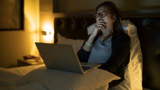 A woman working on her laptop in bed yawns because she is so tired.