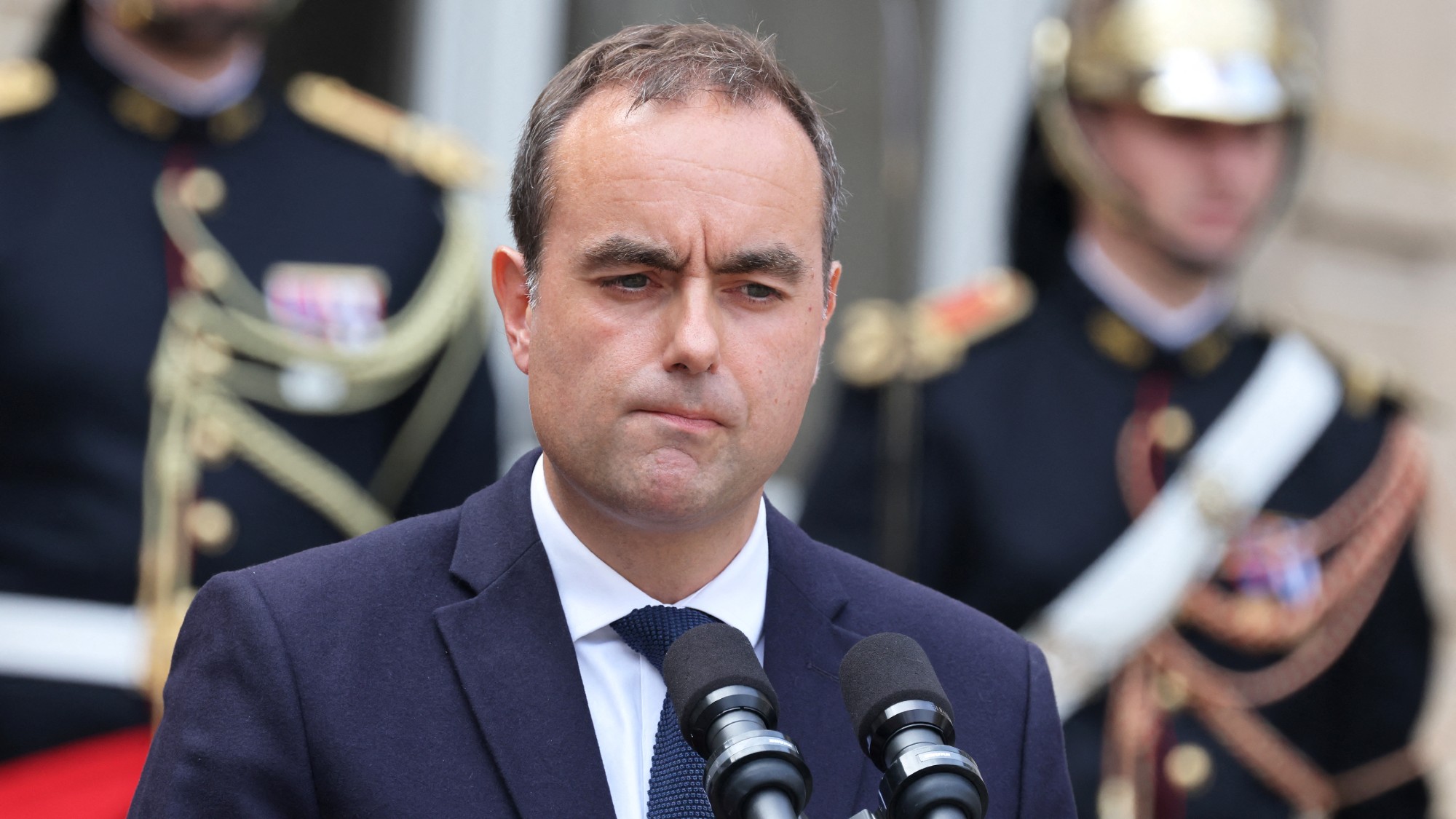 French premier Sebastien Lecornu in a blue suit at a pair of microphones. he looks confused or pensive