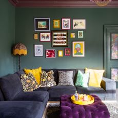 A dark green-painted living room with a dark blue velvet corner sofa and a purple ottoman
