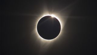 a total solar eclipse with the moon blocking out the sun and only the outer atmosphere of the sun is visible as white light streaming out in all directions.
