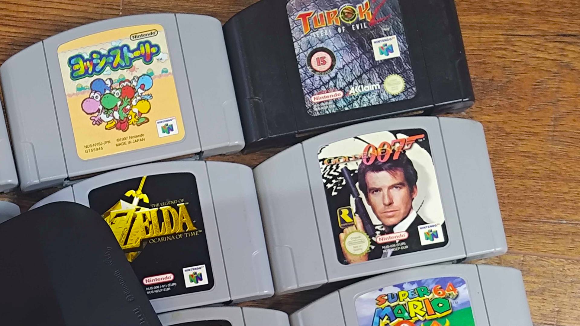 Close up of N64 carts including Yoshi's Story, Zelda Ocarina of Time, Turok 2, and oo7 Goldeneye on woodgrain floor.