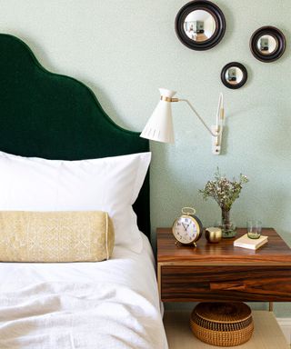 A green wallpapered bedroom with a green velvet headboard and wall mounted bedside lamp