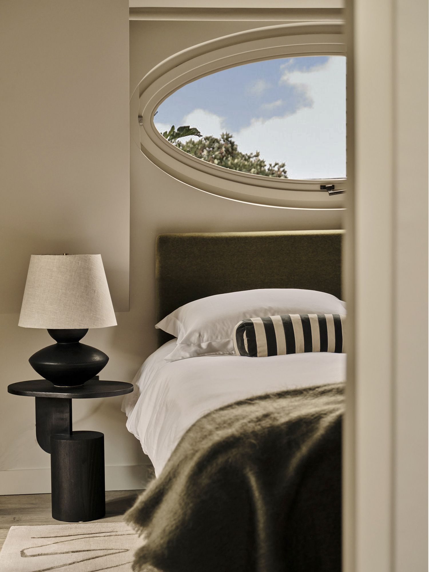 modern bedroom with sculptural side table with table lamp, bed made with white sheets, striped bolster, and green throw, with round window above bed