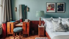 A layered, transitional hotel suite featuring a series of figurative, colorful artworks on its walls, patterned furniture, including rugs, armchairs, and sofas, split-color pillars, and a buttery, tile blue, and green palette.