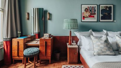 A layered, transitional hotel suite featuring a series of figurative, colorful artworks on its walls, patterned furniture, including rugs, armchairs, and sofas, split-color pillars, and a buttery, tile blue, and green palette.