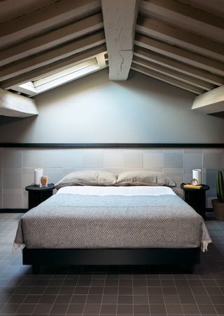 A bedroom with a large bed, black floor tiles, light grey wall tiles, two round bedside tables and a potted cactus.