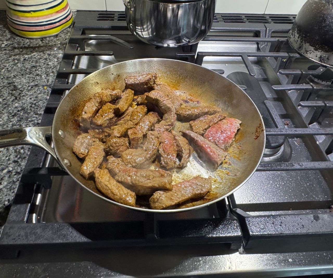 A small stainless steel frying pan on a gas stove filled with browned steak strips