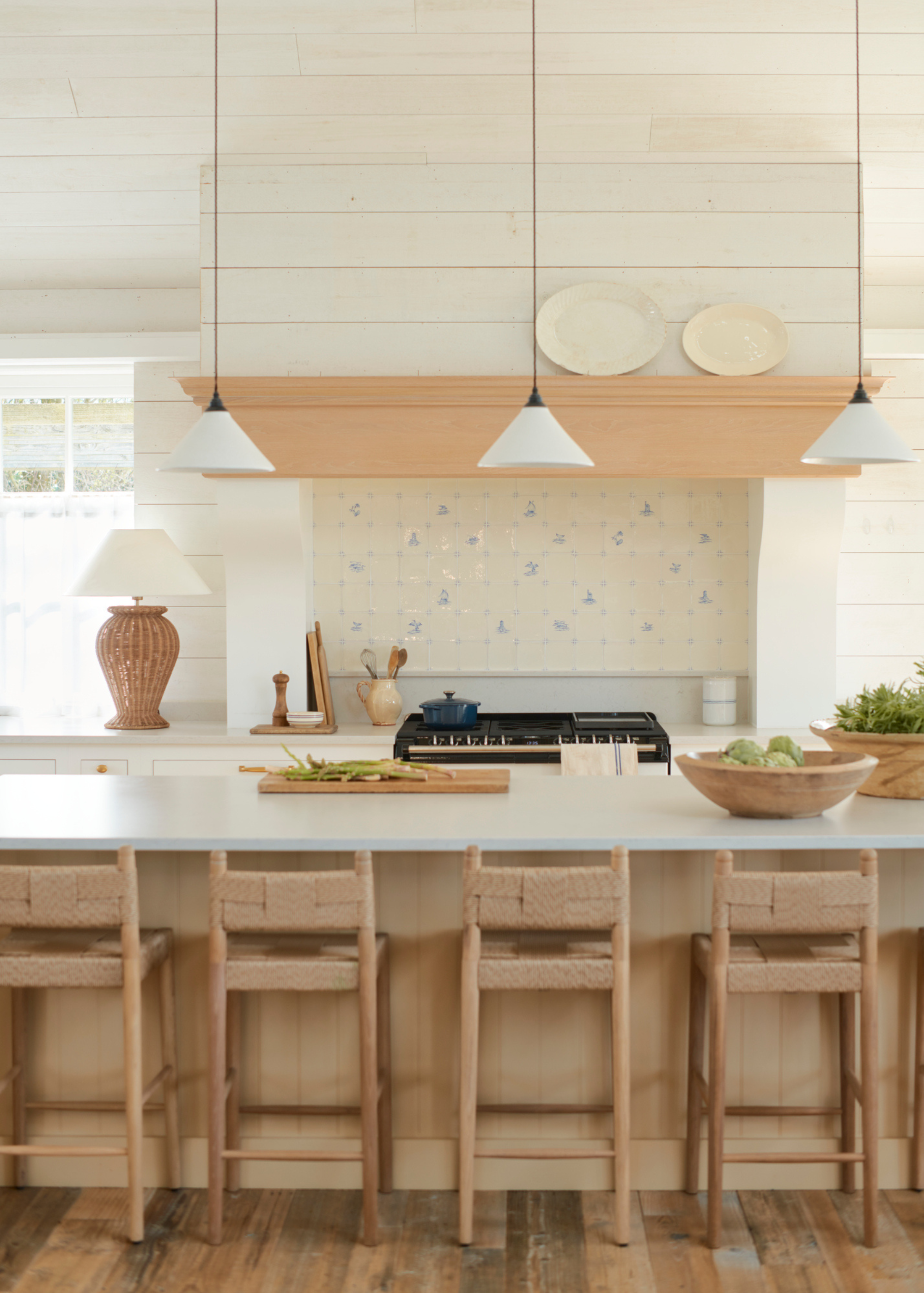 A light wood and off white kitchen with an island in front of a cooker, the backsplash is made up of a stone upstand and delft tiles for added character