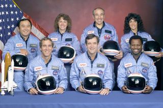 three people sit at the front and four people stand behind them, everyone is smiling and wearing blue NASA jumpsuits.