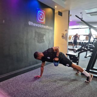 Renel Scarlett performs a kettlebell renegade row inside The Works Health Club gym. His body is elevated off the floor and held in a straight line, supported by his toes and his right hand. His right arm is straight while his left arm is bent and he is rowing a kettlebell up towards his rib cage. In the background we see people on treadmills.