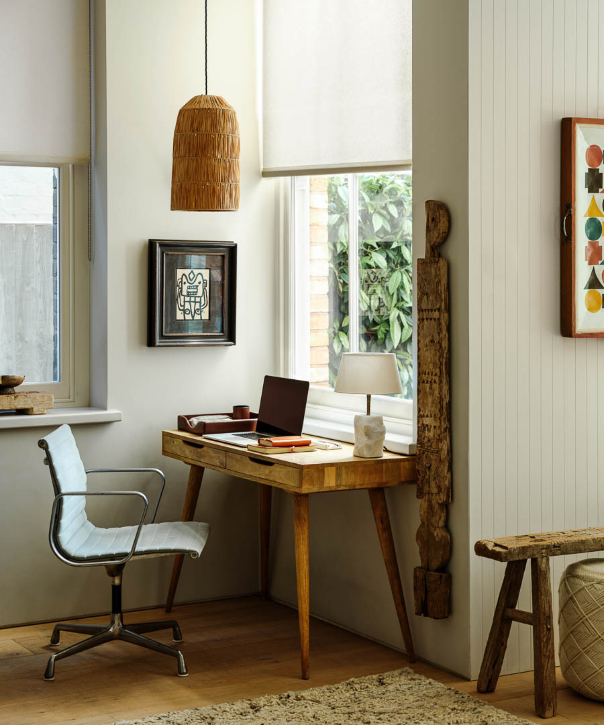 wooden desk placed in front of window with cream roller blind pulled half way down, small white table lamp and rafia pendant hanging above desk and chair
