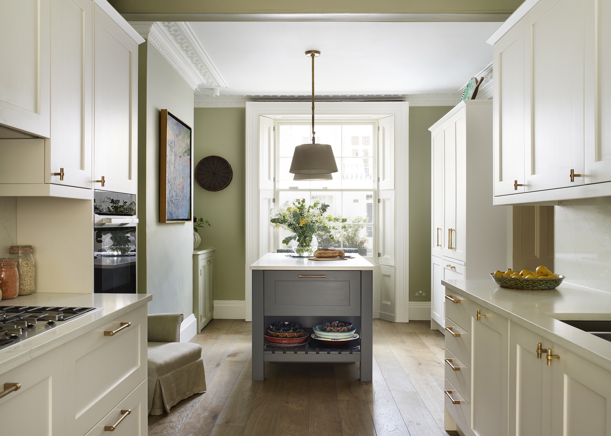 A white and green kitchen with a grey colored island in the middle that has open shelving at the bottom. There are also large pendant lights hanging above the island.