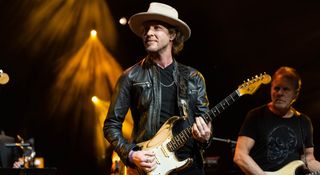 Kenny Wayne Shepherd in wears a wide-brimmed hat and leather jacket onstage and plays a Fender Stratocaster under the stage lights.