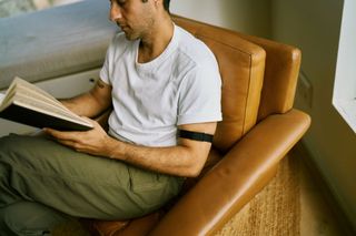 A man sits in a leather armchair reading a book. A Visible armband can be seen on his upper arm.