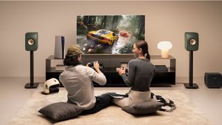 KEF Wireless II speakers on either side of a TV while a couple on cushions play video games