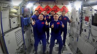 six astronauts in blue flight suits give a thumbs-up aboard a white-walled space station