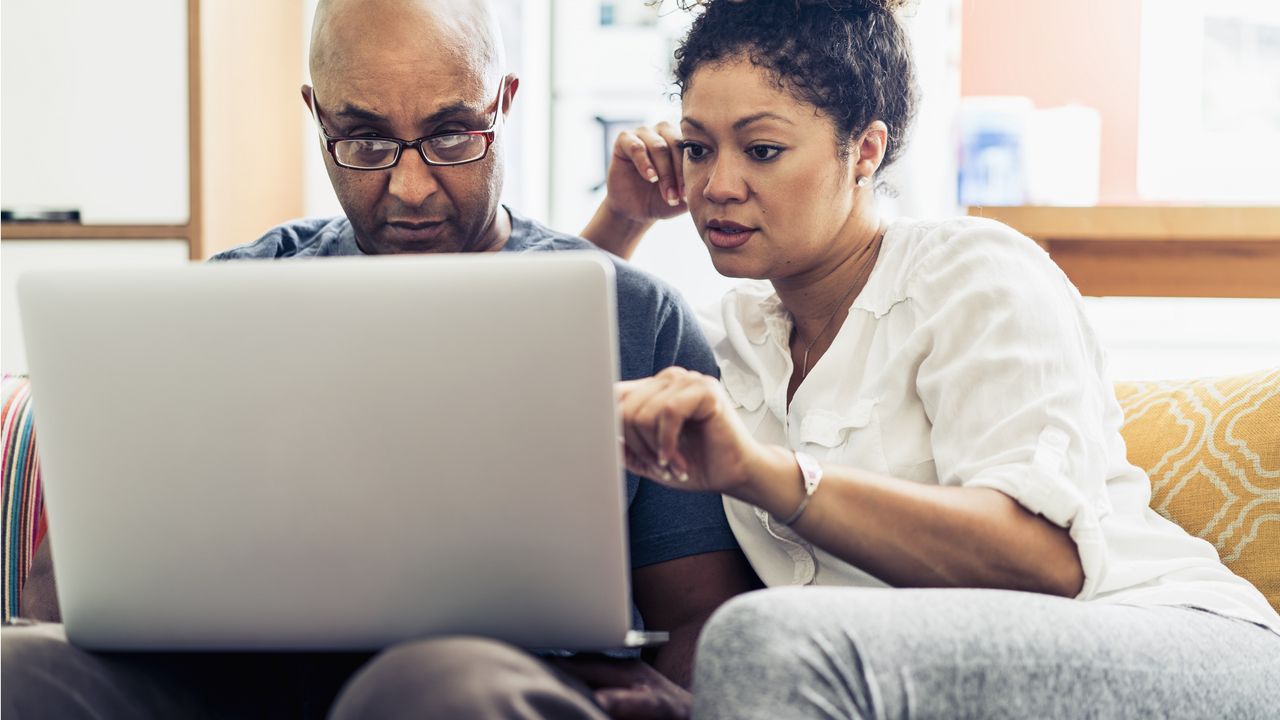 A married couple sit next to each other on a sofa and look at the screen of a laptop on the husband&#039;s lap.