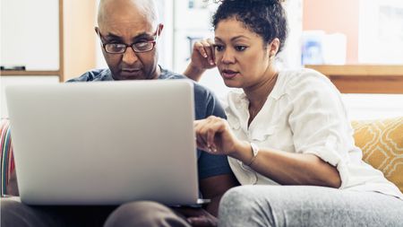 A married couple sit next to each other on a sofa and look at the screen of a laptop on the husband's lap.