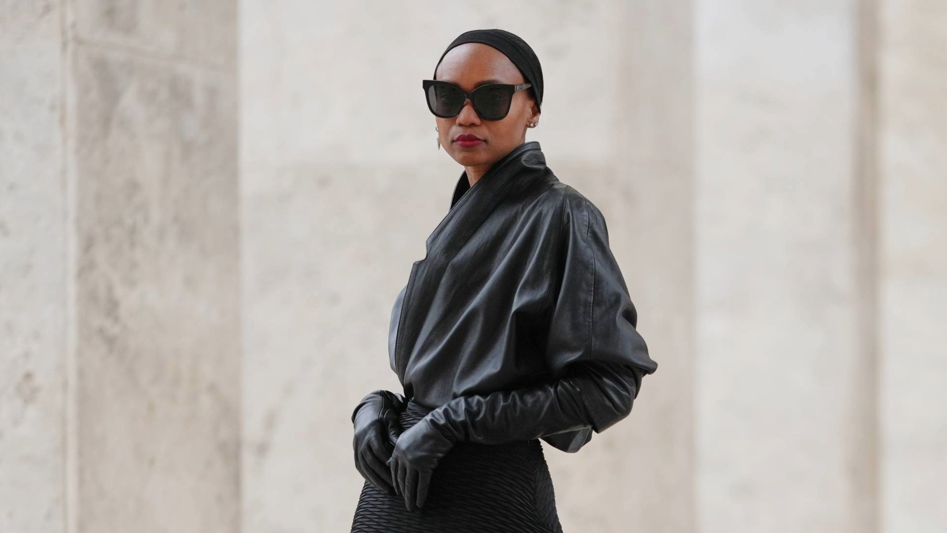 PARIS, FRANCE - DECEMBER 20: Emilie Joseph wears slicked-back hair held with a minimalist black headband, natural makeup with a smooth complexion and defined lips, black rectangular sunglasses, a vintage black leather jacket styled as a wrap top with a cinched waist creating a sculptural line, a black asymmetric pleated skirt by COS layered over black leggings, vintage black leather gloves, black leather slingback heels shoes with a square silver buckle by Saint Laurent, during a street style fashion photo session, on December 20, 2025 in Paris, France. (Photo by Edward Berthelot/Getty Images)
