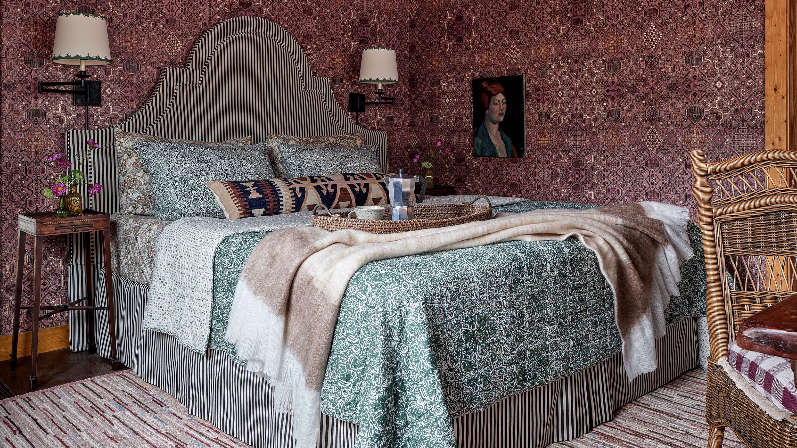 a primary bedroom with purple printed wallpaper, a large striped upholstered bed with headboard and skirt, antique furniture and a striped rug